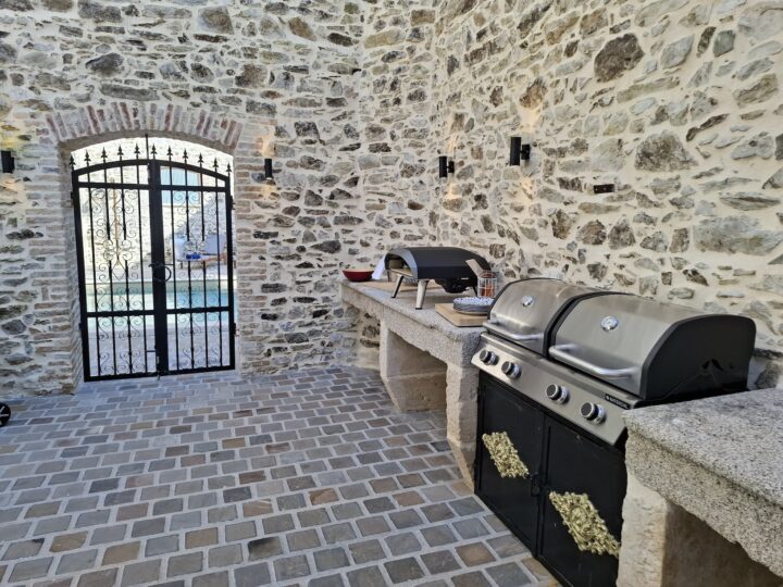 Outdoor kitchen with stone walls and floor, featuring a grill, a pizza oven, worktops, and a black wrought iron gate leading outside. Cooking utensils and bowls are arranged on the counter.