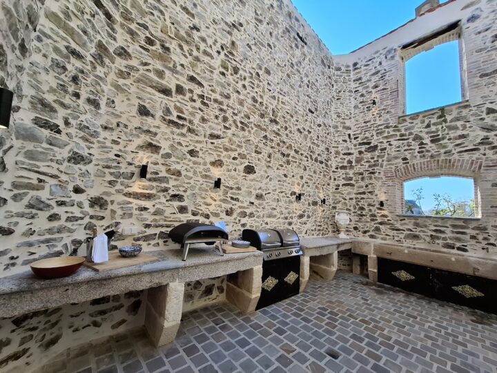 Outdoor kitchen with stone walls, a built-in worktop, pizza ovens, a barbecue, and some kitchen items. Large open windows and sky are visible above, giving an airy and rustic atmosphere.