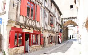 A narrow cobbled street lined with old timber-framed buildings; one has red shutters and a shopfront. In the background, a stone archway crosses over the street. Two people walk in the distance.