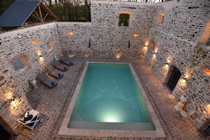 A rectangular swimming pool is surrounded by stone walls and lit wall sconces. Loungers line one side, and small tables and chairs are placed in the corners. The area has a rustic, courtyard feel with a brick floor.