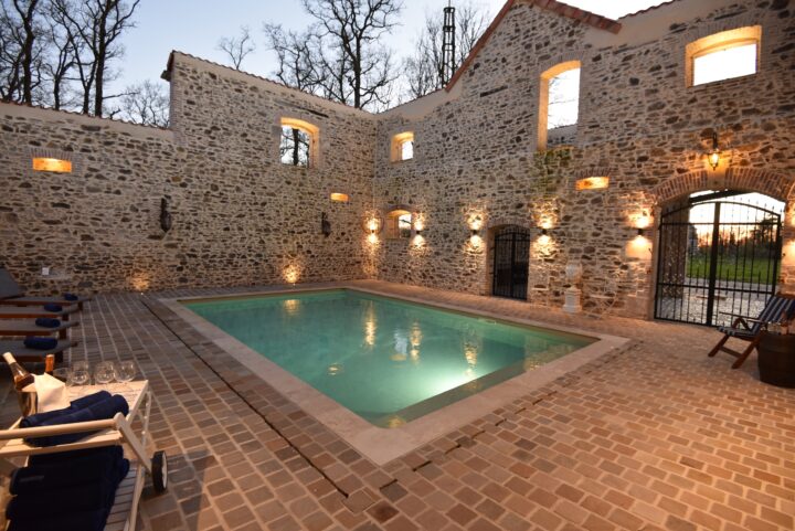 An indoor pool surrounded by rustic stone walls with arched windows and warm lighting, lounge chairs with towels and wine glasses nearby, creating a cosy, inviting atmosphere in the evening.