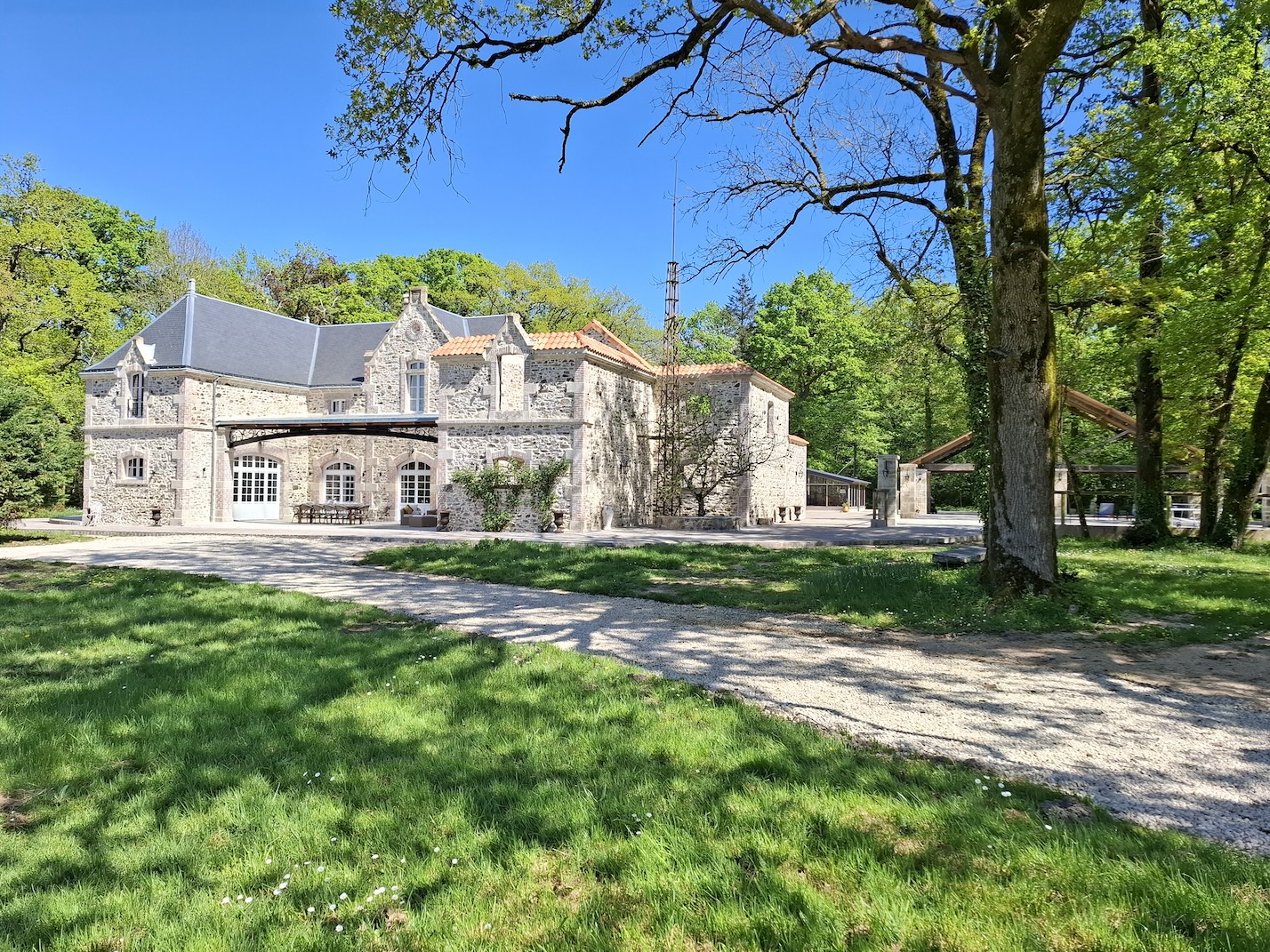 A stone building with arched windows and a grey roof sits surrounded by green grass, trees, and a gravel drive under a clear blue sky.