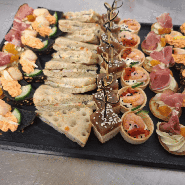 A platter of assorted bite-sized appetisers, including savoury tarts, pitta slices, bread triangles with sesame seeds, prawns, smoked salmon, cucumber, whipped toppings, and cured meats, arranged neatly on a black slate tray.