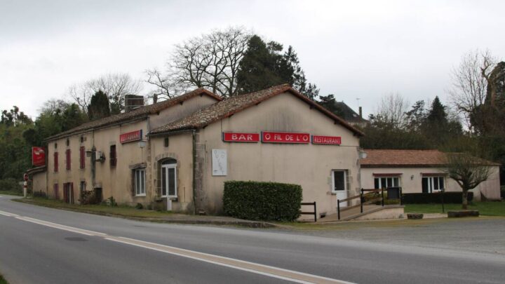 A rustic building with beige walls and red roof sits beside a road, featuring red signs labelled BAR and RESTAURANT. Trees and bushes surround the structure under an overcast sky.