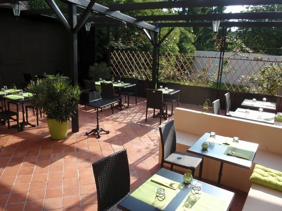 Outdoor restaurant patio with terracotta tile floor, black tables and chairs, green potted plants, and green serviettes on tables. Sunlight brightens the space, and a wooden trellis provides partial privacy.