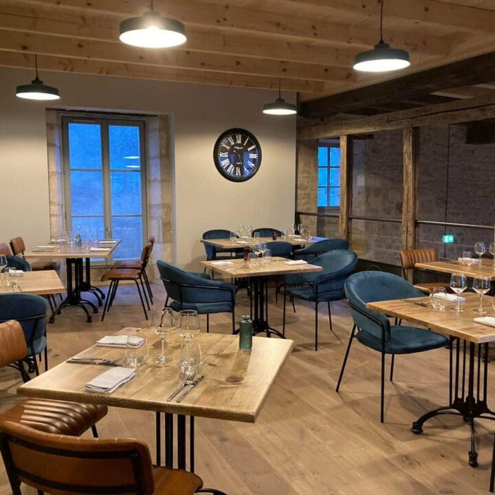 Modern restaurant interior with wooden floors, exposed beams, and large windows. Tables are set with glasses and serviettes, surrounded by brown leather and blue velvet chairs. A large wall clock decorates the space.