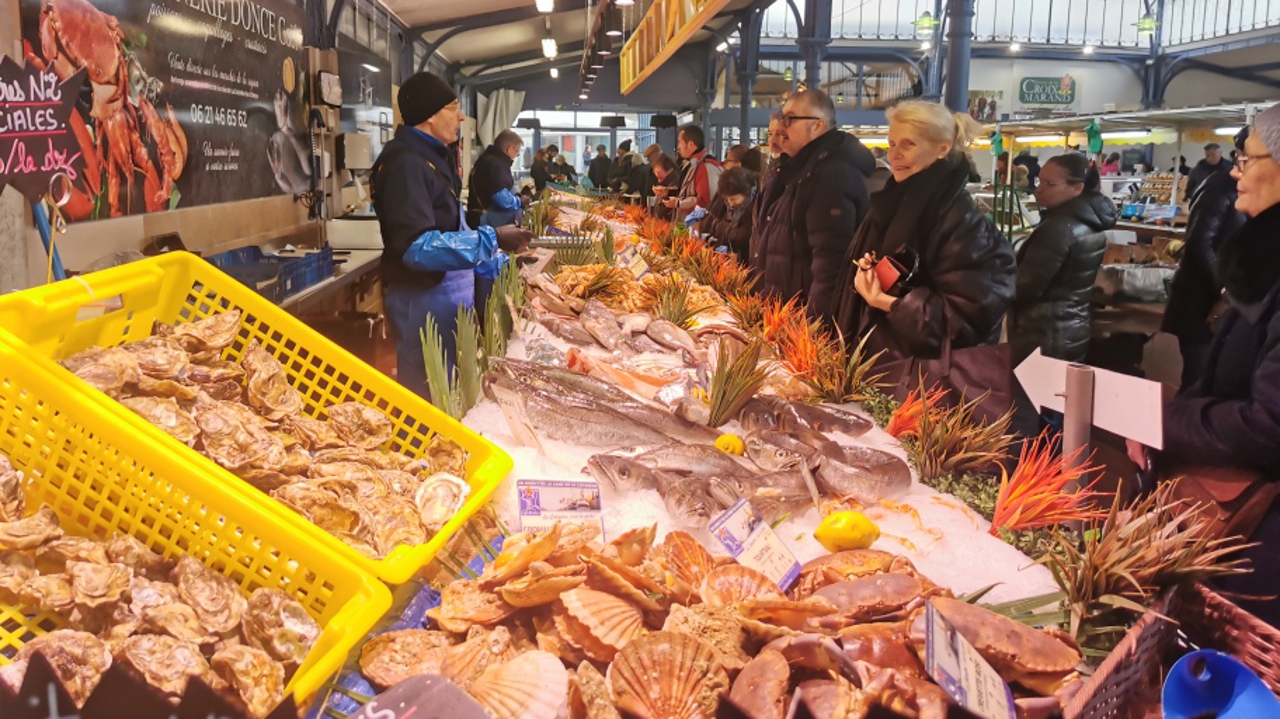 A busy indoor seafood market with vendors selling fresh fish, oysters, and shellfish on ice to customers standing at the counter. Bright yellow baskets hold oysters, and various sea creatures line the display.