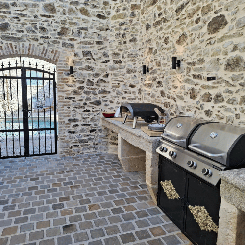 Outdoor stone patio with a gated entry, built-in barbecue, pizza oven, and worktop on a cobblestone floor, surrounded by high stone walls.