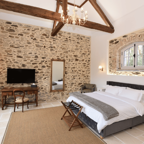 A cosy bedroom with a stone feature wall, exposed wooden beams, a double bed with white linen, a chandelier, a TV on a wooden desk, a mirror, and a window with a stone frame.