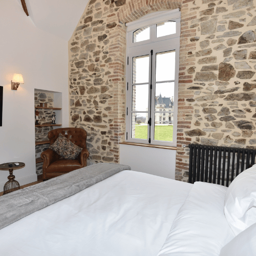 Cosy bedroom with a stone feature wall, large window overlooking a lawn and distant building, brown armchair, bookcase, side table, and white bedding.