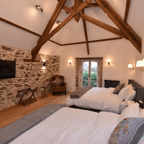 A cosy bedroom with two double beds, exposed wooden beams, a stone feature wall, a TV, an armchair, and French doors leading to a balcony with a view outside. Warm lighting completes the inviting atmosphere.