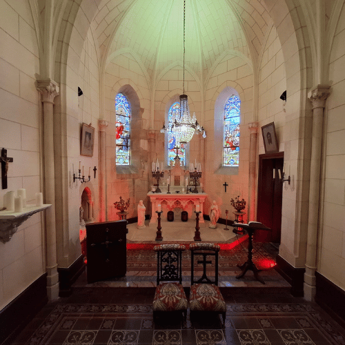 A small, ornate chapel with a chandelier, stained glass windows, candles, religious paintings, and an altar, viewed from the entrance. Two upholstered chairs face the altar on patterned tiled flooring.