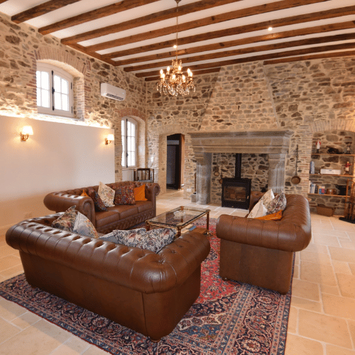 Rustic living room with stone walls, wooden ceiling beams, leather sofas, a chandelier, a fireplace, and a patterned rug on a light stone floor.