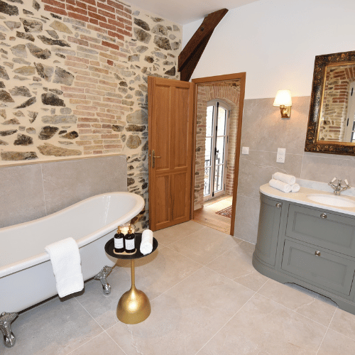 A rustic bathroom with a clawfoot bath, rolled towels, soaps on a small table, a grey vanity unit, a mirror, stone and brick walls, and an open wooden door leading to a hallway.