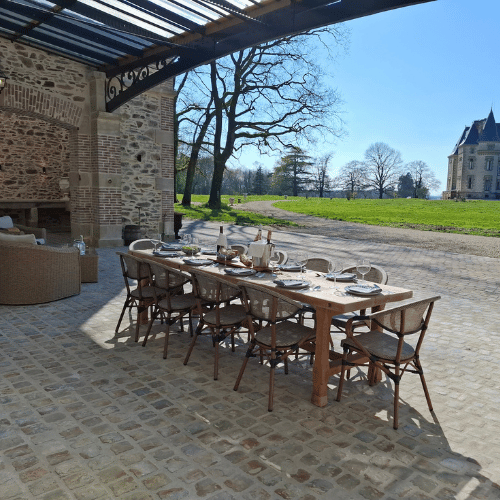 A long wooden dining table with chairs is set for a meal on a cobbled patio, next to a brick building. In the background, there is a grassy area, trees, and a distant castle-like structure.