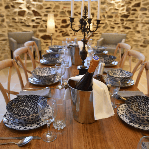 A wooden dining table set with blue-patterned crockery, glasses, and cutlery. Two bottles in an ice bucket and a black candelabrum serve as the centrepiece. Stone wall and chairs in the background.