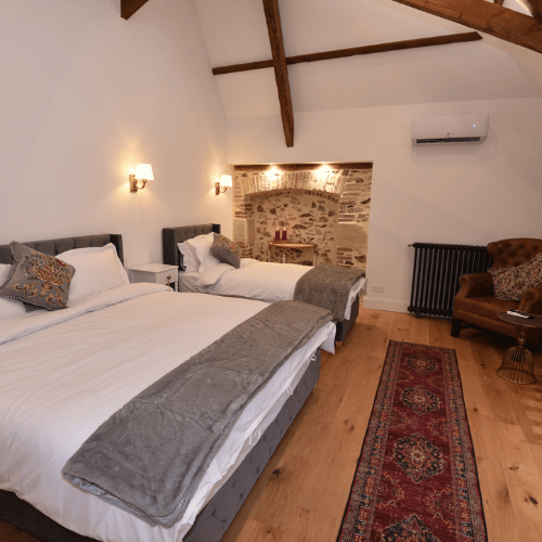 A cosy bedroom with exposed beams, one double and one single bed, grey throws, patterned cushions, an armchair, a red rug, and a stone feature wall with shelves and decorative items.