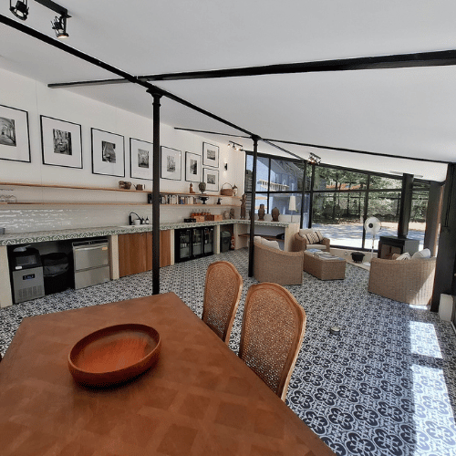 Modern open-plan kitchen and living area with tiled floor, glass walls, wooden dining table, wicker chairs, lounge seating, and framed photos on the white wall, filled with natural sunlight.