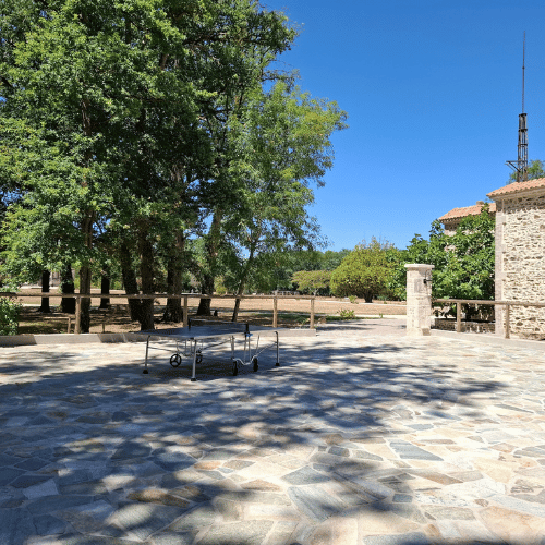 A stone patio with a folded table tennis table sits in the sun. Trees provide shade in the background, and part of a stone building with a tiled roof is visible on the right. The sky is clear and blue.