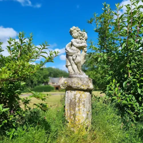 Weathered stone statue of two children on a plinth, surrounded by lush green foliage and grass, with a blue sky and distant house in the background.