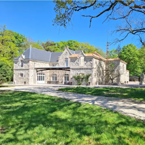 Large stone house with arched windows and a sloping roof, surrounded by green grass, trees, and a gravel drive under a clear blue sky.