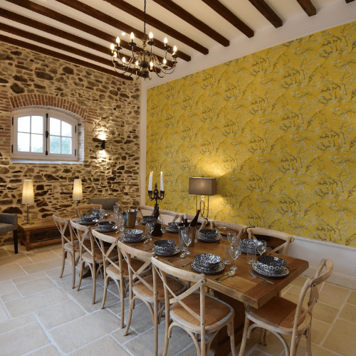 A rustic dining room with a long wooden table set for ten, stone walls, yellow floral wallpaper, wooden beams on the ceiling, and a chandelier overhead. A window lets in natural light.