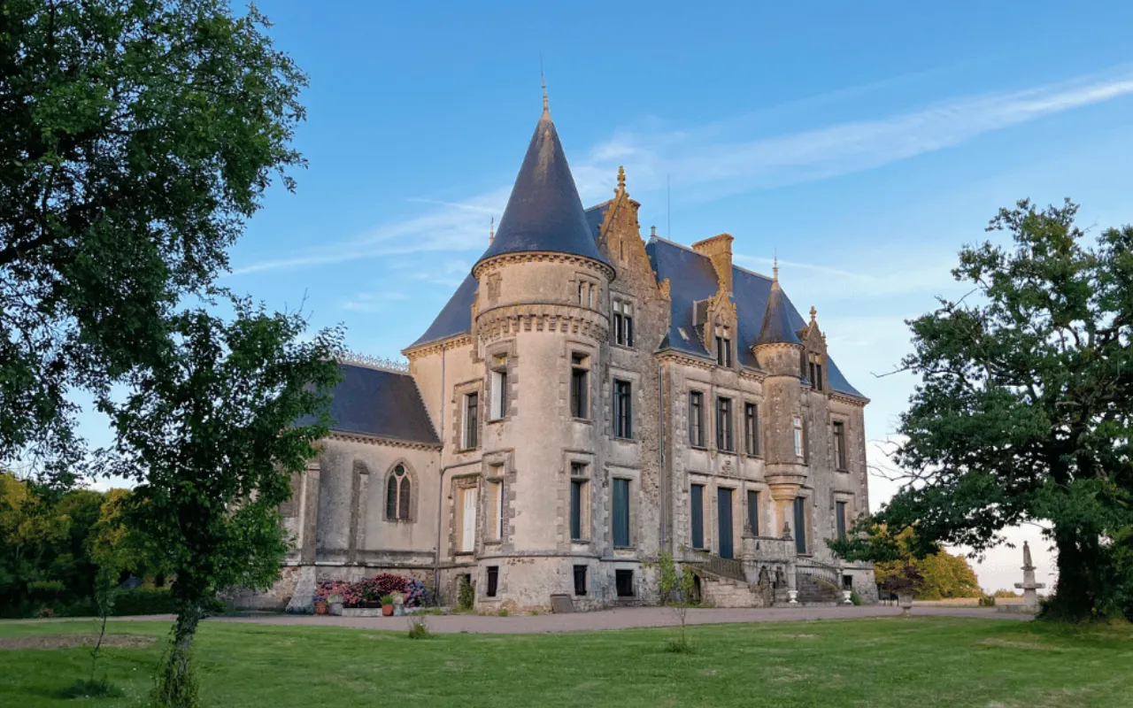 A large stone château with pointed towers and arched windows stands on a grassy lawn with trees, under a clear blue sky.