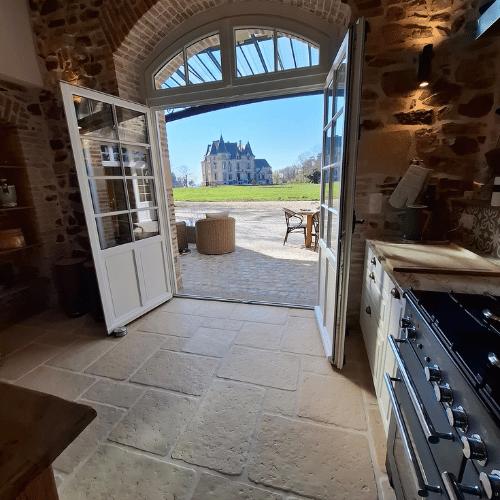 View from a rustic kitchen with stone walls and open glass doors leading to a sunny patio; a grand château is visible in the distance across a courtyard.