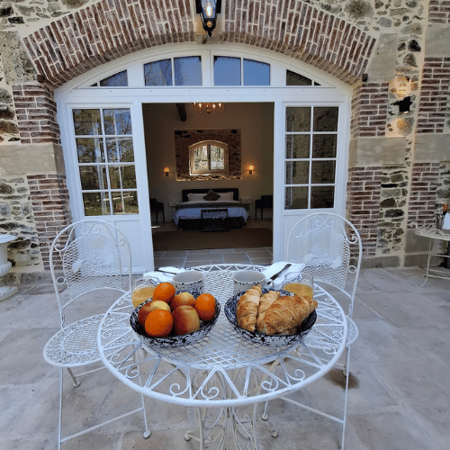 A white metal table with fruit and pastries sits on a patio in front of a stone building with large arched glass doors, revealing a bedroom inside.