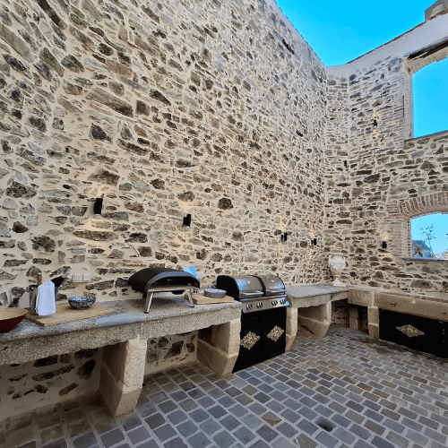 Outdoor stone kitchen with pizza oven, grill, and sink built into a rustic stone wall; open ceiling and bright blue sky above.