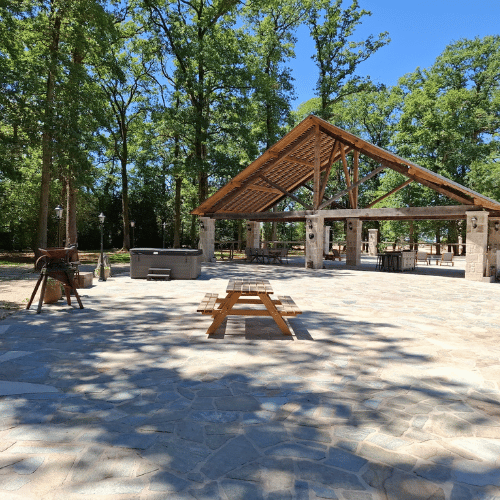 A wooden picnic table sits on a stone patio under a clear sky, with a pavilion and trees in the background. A barbecue and rubbish bin are visible to the left.