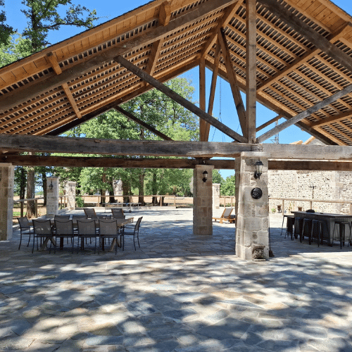 Open-air pavilion with a wooden roof, stone pillars, and tiled floor. There are tables and chairs arranged under the structure, surrounded by trees and natural light on a sunny day.