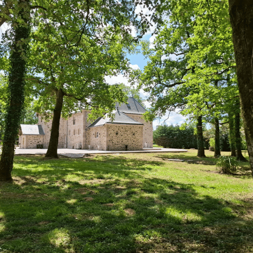A stone building with a grey roof is surrounded by green trees and grass on a sunny day, with light filtering through the leaves.