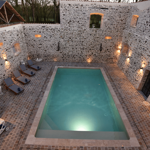 Rectangular swimming pool surrounded by stone walls, with five loungers on one side and evening lights illuminating the stone, creating a cosy courtyard atmosphere.