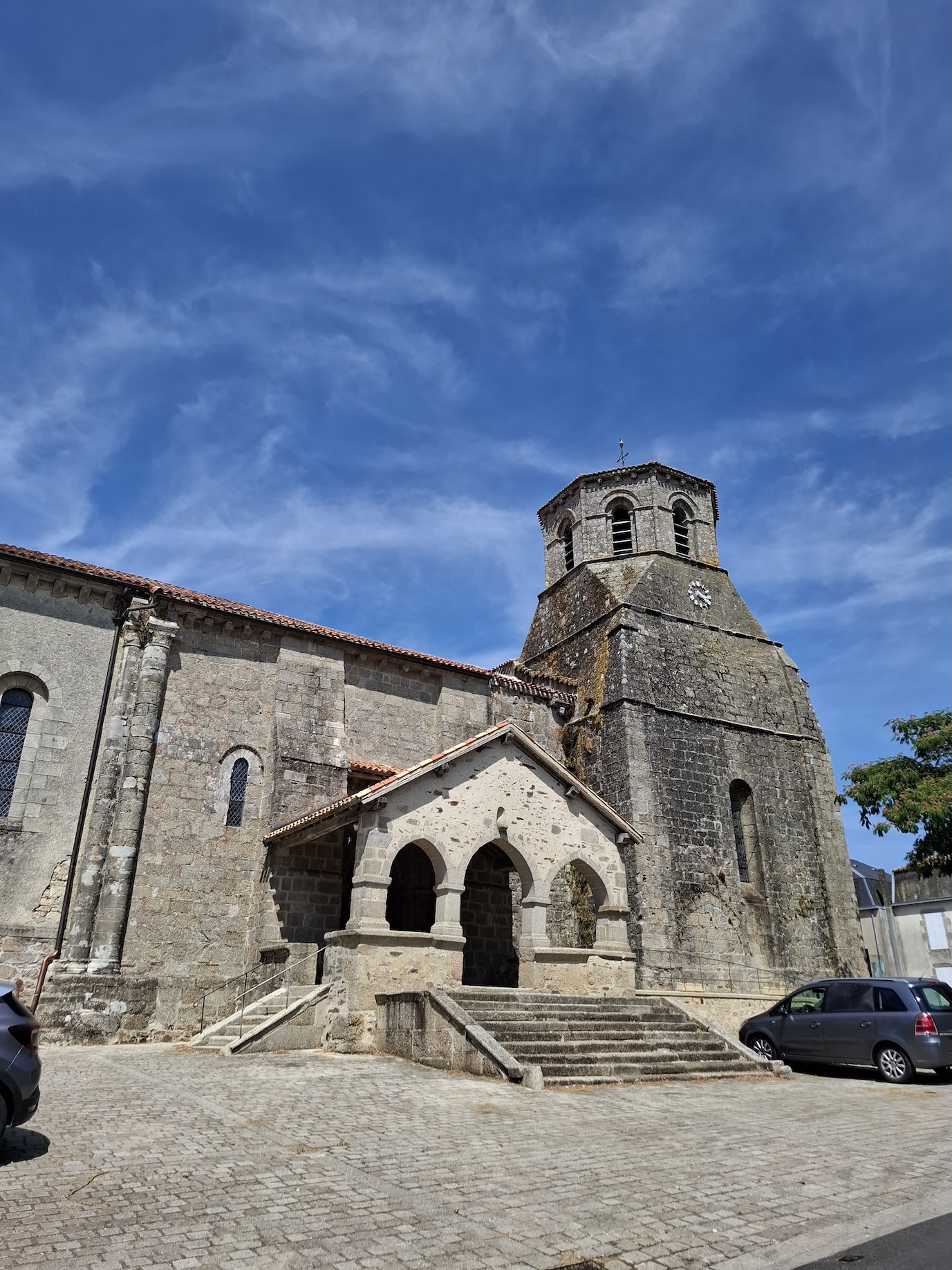 Stone church with a square bell tower and arched entrance under a bright blue sky. Two parked cars and a cobbled courtyard are visible in the foreground.