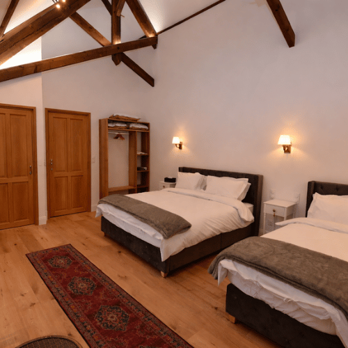 A cosy bedroom with two beds, wooden floors, exposed ceiling beams, wall lights, an open wardrobe, and a red patterned rug across the room.