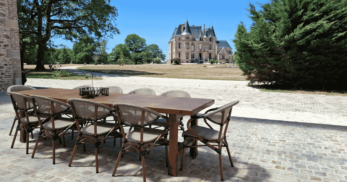 A long wooden outdoor dining table with ten chairs sits on a stone terrace, overlooking a large grassy area and a grand château in the background under a clear blue sky.