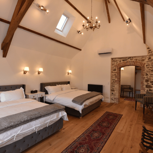 A cosy bedroom with two double beds, wooden floor, exposed beams, wall sconces, chandelier, and a brick archway leading to another room. A skylight and rug add warmth to the space.
