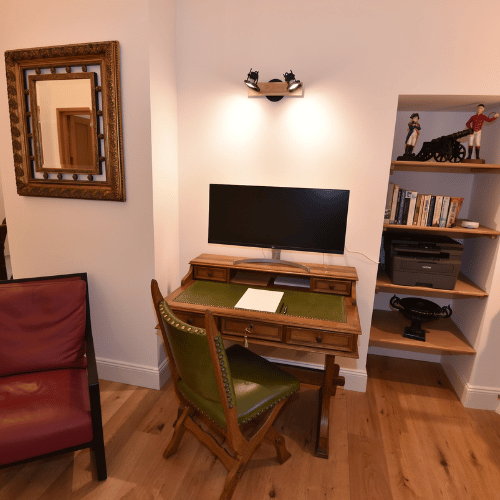 A small home office features a wooden desk with a green chair, a monitor, and a notebook. A bookcase with books and figurines stands to the right, and a framed mirror hangs on the left wall above a red chair.