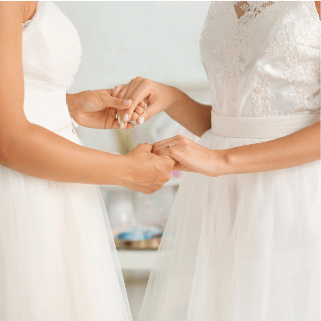 Two women in white wedding dresses hold hands, facing each other. Only their torsos are visible, with delicate lace and tulle details on their dresses. The background is softly blurred.