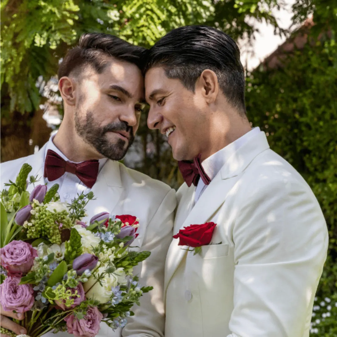 Two men in white suits and red bow ties embrace and smile, holding a bouquet of flowers. They appear happy and are surrounded by greenery outdoors, suggesting a wedding or special occasion.