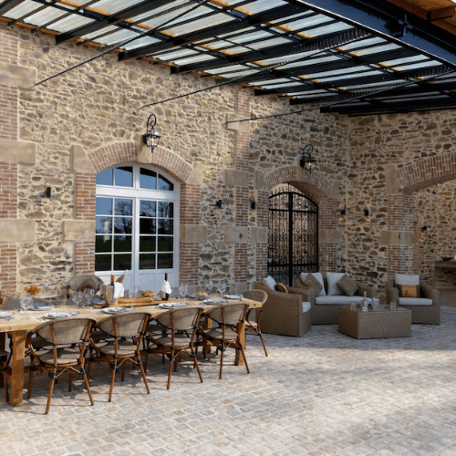 Outdoor patio with stone walls, a long wooden dining table set with chairs, and a seating area with sofas and a coffee table. The space is covered by a glass roof and lit by wall-mounted lanterns.