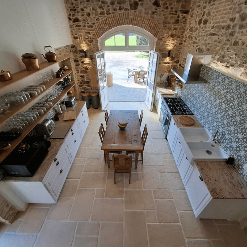 A bright rustic kitchen with stone walls, white cupboards, open shelves, a wooden table and chairs, patterned splashback, and open double doors leading outside.