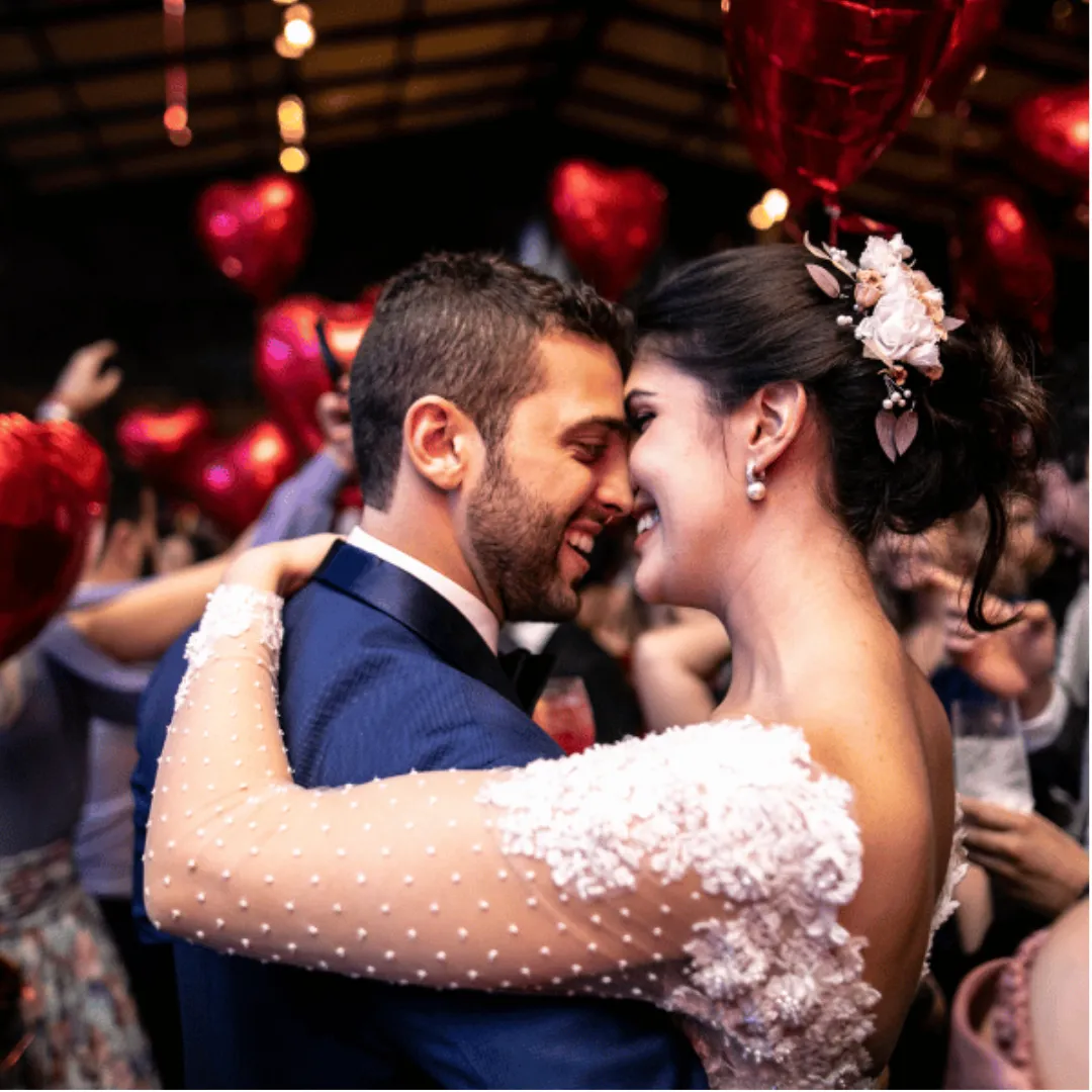A bride and groom smile and embrace while dancing, surrounded by guests holding red heart-shaped balloons at a festive wedding reception.