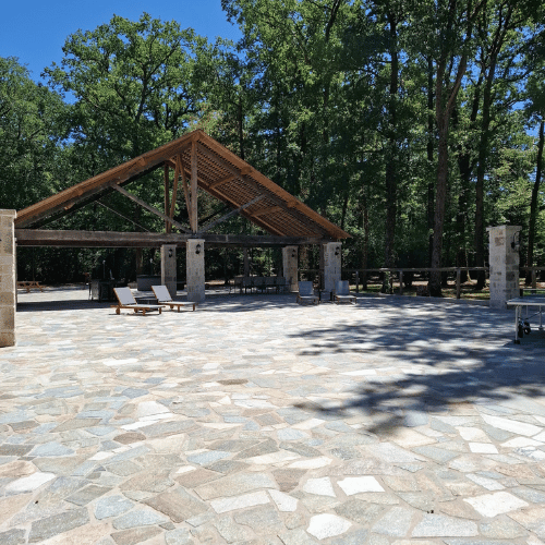 Open stone patio with a wooden-roofed pavilion, lounge chairs, and tall trees in the background under a clear blue sky.