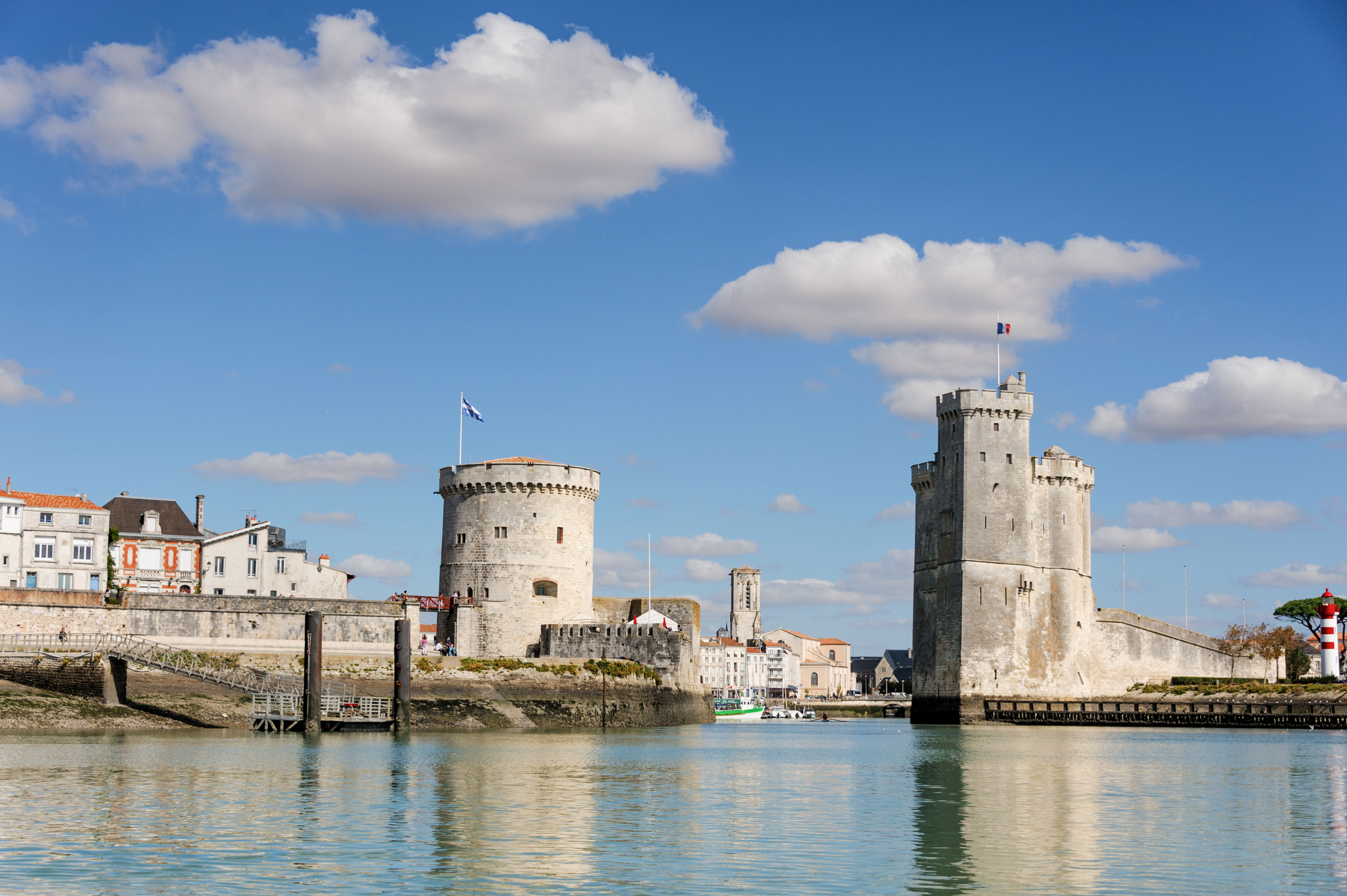 Two historic stone towers stand by the water at a harbour entrance under a blue sky with scattered clouds, with buildings and a lighthouse in the background.