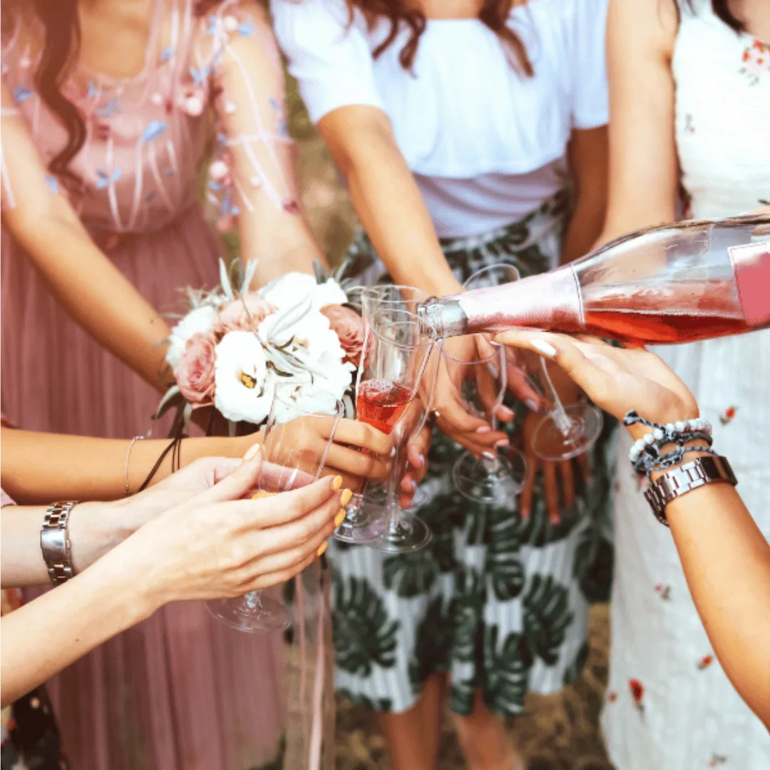 A group of women clink glasses as one pours rosé wine. Hands hold floral bouquets and wear bracelets, suggesting a celebration or special occasion outdoors.