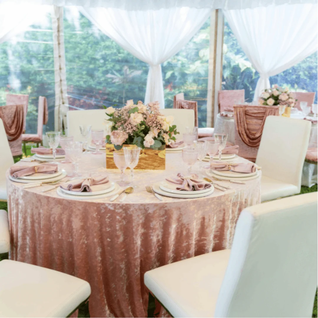 A round table with a pink velvet tablecloth, white chairs, pink serviettes, gold centrepiece with flowers, glassware, and place settings is set for an elegant event in a marquee with white drapes.