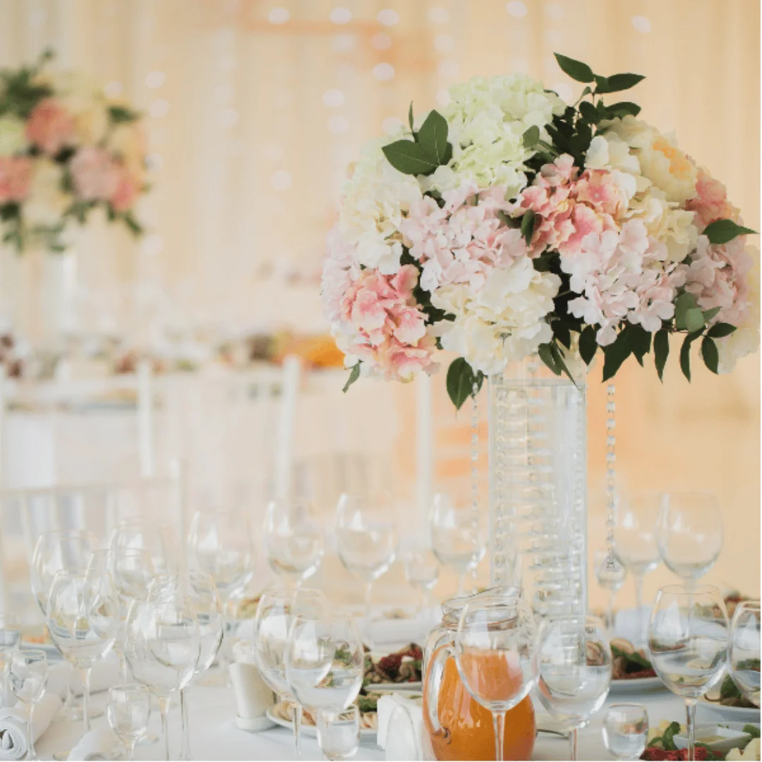 Elegant wedding table setting with glassware, plates, and food, featuring a tall centrepiece of pink and white hydrangeas and greenery in a clear vase. Soft, warm lighting and blurred background with similar decor.