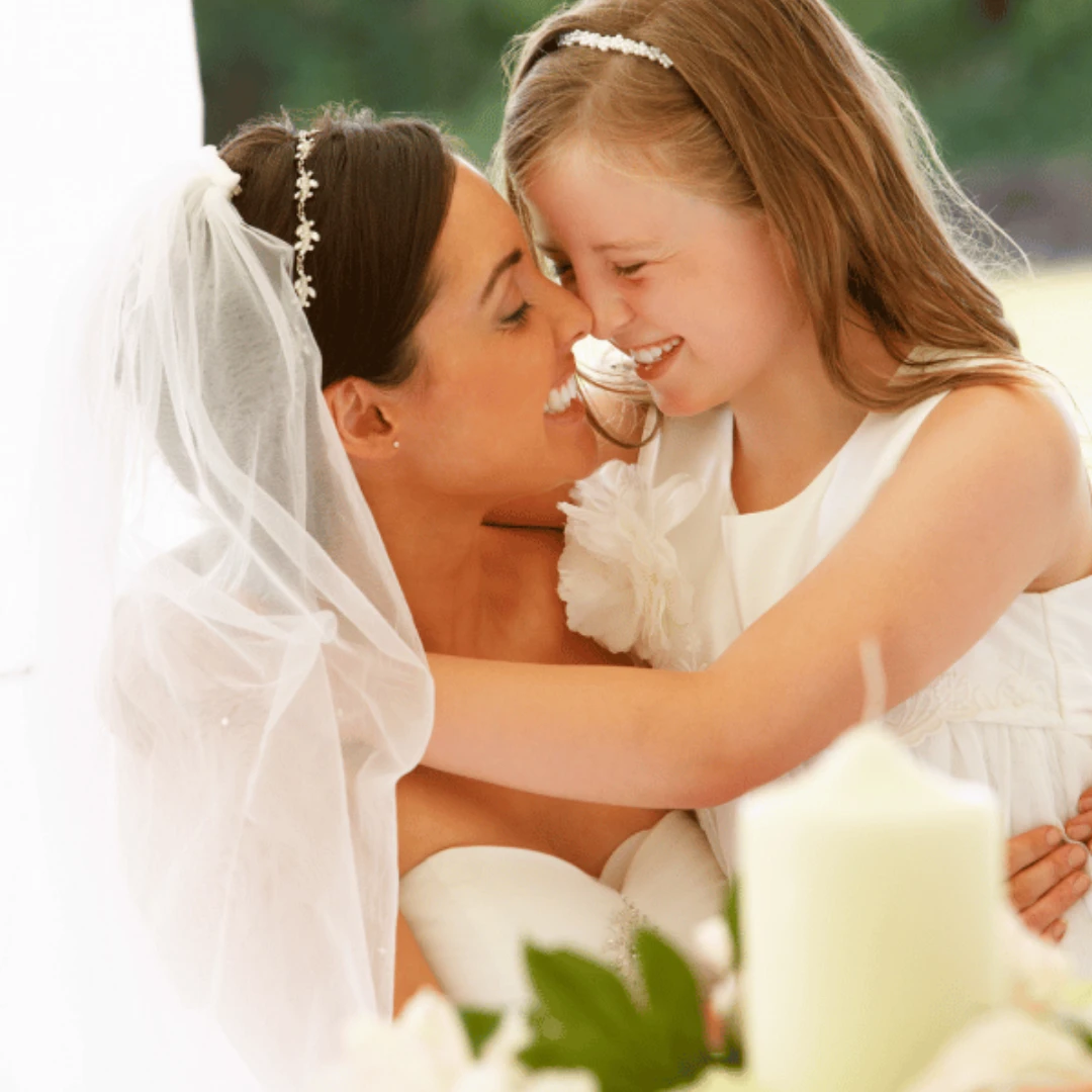 A bride in a veil hugs and smiles lovingly with a young girl in a white dress, both touching noses and appearing joyful.